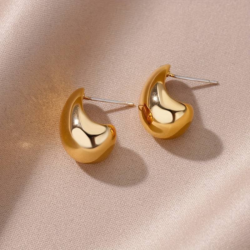 Displayed on a soft, beige fabric with a subtle sheen, the Water Drop Curved Shaped Stud Earrings glisten with their smooth, glossy surfaces. The small, gold earrings boast an elegant and fluid design that casts gentle shadows on the fabric.