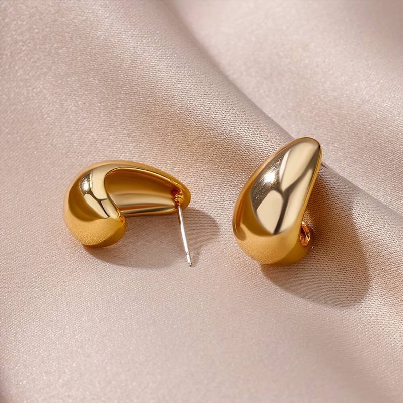 A pair of Water Drop Curved Shaped Stud Earrings with a gold, smooth, glossy finish are artfully arranged on an elegant, cream-colored, silky fabric background. One earring lies on its side revealing the post, while the other stands upright.