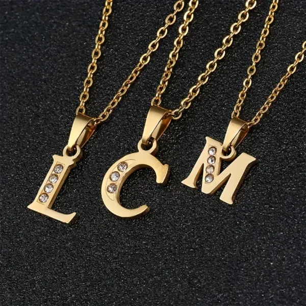 Three gold necklaces, each featuring a pendant in the shape of a letter—"L," "C," and "M"—reminiscent of the Stainless Steel Letter Necklace, are adorned with small, glittering diamonds. The necklaces are elegantly displayed on a black, textured surface.