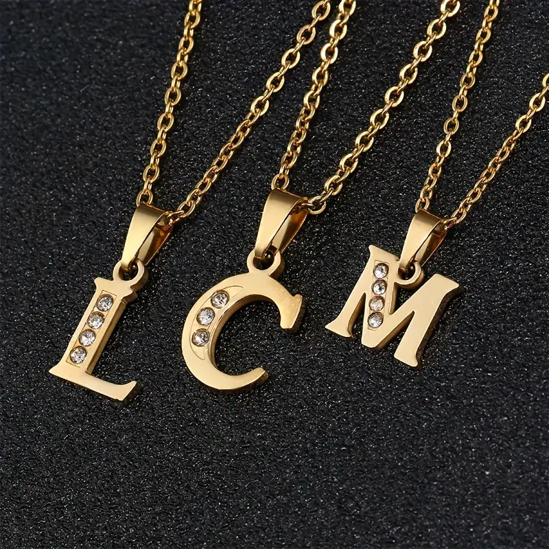 Three gold necklaces, each featuring a pendant in the shape of a letter—”L,” “C,” and “M”—reminiscent of the Stainless Steel Letter Necklace, are adorned with small, glittering diamonds. The necklaces are elegantly displayed on a black, textured surface.