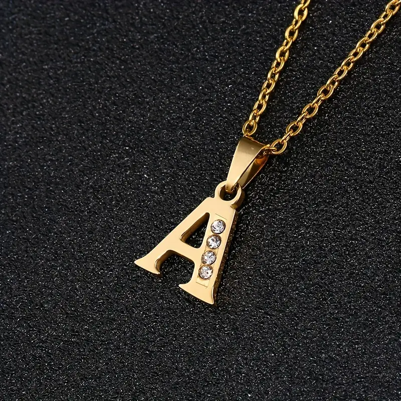 Presenting the Stainless Steel Letter Necklace: a stunning gold necklace featuring a pendant in the shape of the letter “A.” It is beautifully adorned with four small diamonds on its right leg and displayed on a black textured background. This elegant piece exudes timeless sophistication, making it a perfect addition to any collection.