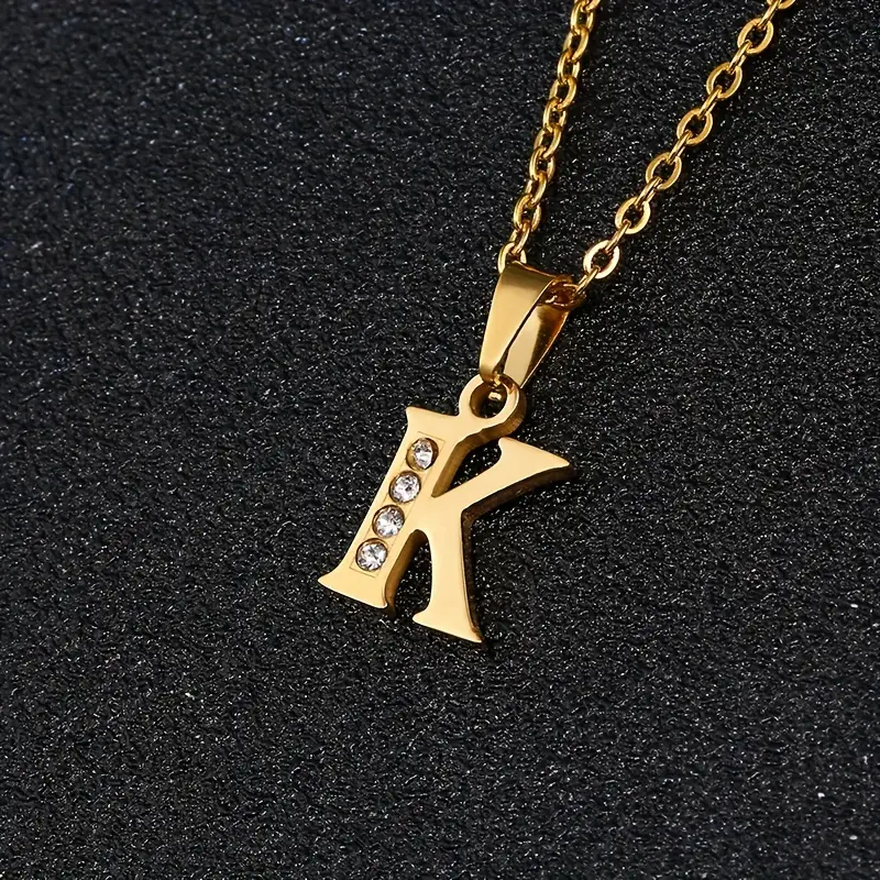 A gold chain necklace featuring a pendant in the shape of the letter “K” from our Stainless Steel Letter Necklace collection. The stainless steel letter is elegantly embellished with four small, sparkling clear crystals arranged vertically along its left side, and it is showcased against a textured black background.