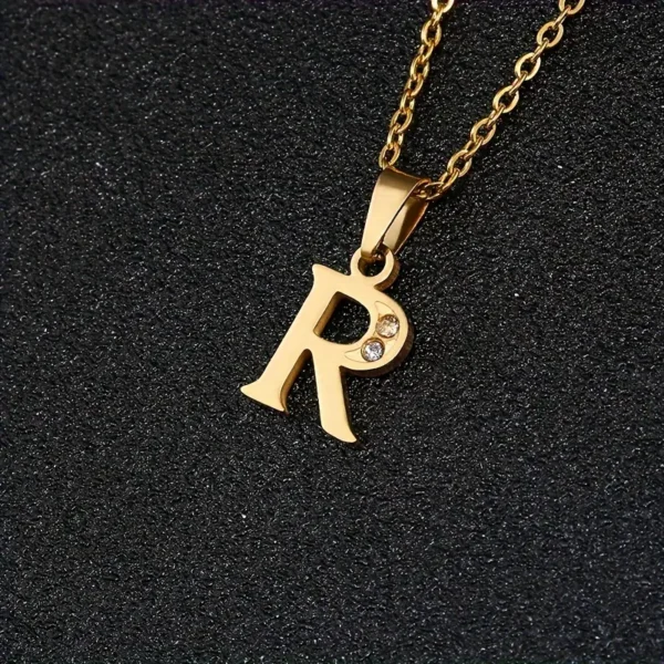 The Stainless Steel Letter Necklace, with a gold pendant shaped like an "R" and adorned with a small sparkling gem in the center, is displayed against a black textured background.