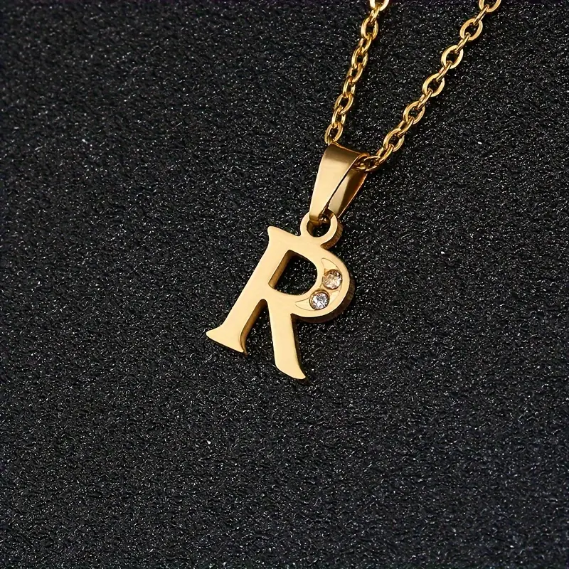 The Stainless Steel Letter Necklace, with a gold pendant shaped like an “R” and adorned with a small sparkling gem in the center, is displayed against a black textured background.