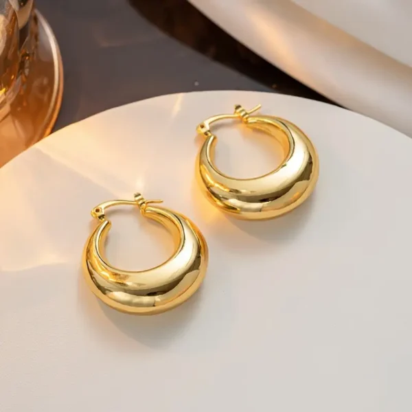 A pair of Elegant French Retro 14K Gold Plated Chunky Hoop Earrings – Minimalist Vintage Style, featuring a thick, round shape, are placed on a smooth, light-colored surface. The background includes soft fabric and subtle warm lighting, highlighting the elegance of these exquisite hoops.