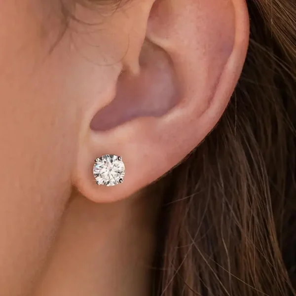 Close-up of a person's ear adorned with Brilliant Round Cut Moissanite Earrings, their brilliance and clarity emphasized by the light. The four-prong design secures the moissanite gem perfectly. The person's hair is partially visible, tucked behind the ear.