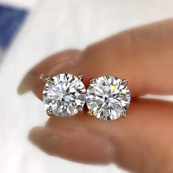 Close-up of two Brilliant Round Cut Moissanite Earrings held between a person's fingers. The four-prong setting allows the brilliance of the moissanite stones to be prominently displayed. The background is softly blurred, highlighting the elegance of these stunning accessories.