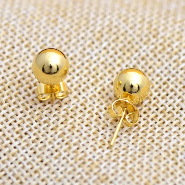 A pair of Stylish 14K Gold Plated Ball Stud Earrings - Perfect Gift for Women and Girls is displayed on a textured beige fabric. One earring lies flat, showcasing the front of the ball stud earring, while the other is angled to reveal its back and post. The earrings have a shiny, reflective finish.