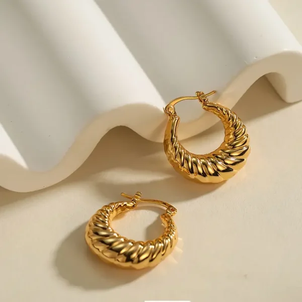 A pair of Delicate Golden Horn Bag Hoop Earrings – Vintage Copper Jewelry for Daily Casual Elegance are displayed on a cream-colored, wavy textured surface. One earring lies flat, while the other stands tall, showcasing their intricate detail and vintage elegance.