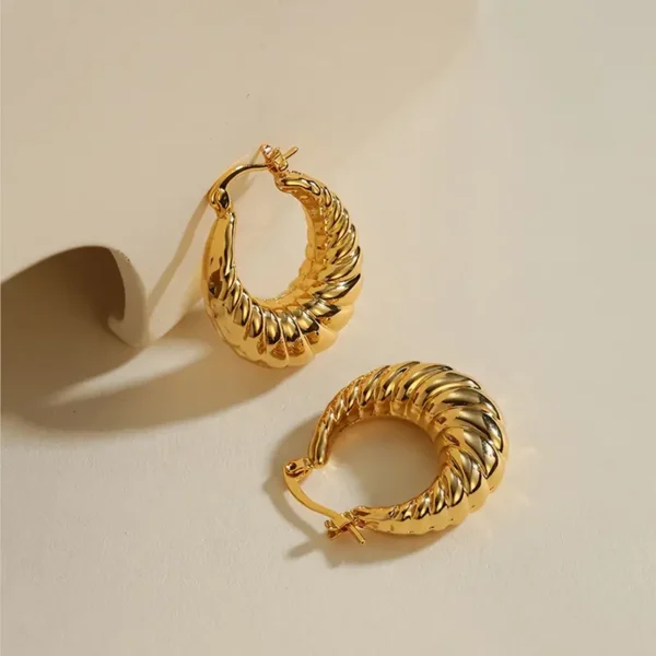 A pair of Delicate Golden Horn Bag Hoop Earrings – Vintage Copper Jewelry for Daily Casual Elegance are displayed on a beige surface. One earring leans against a curved object, while the other lies flat. The lighting highlights their glossy, polished finish, perfectly capturing the elegant casual style.