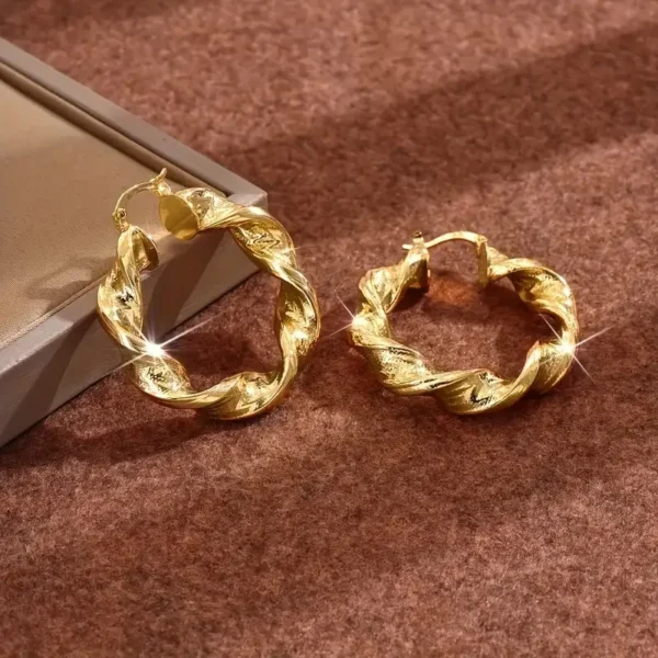 A pair of Twisted Rope Golden Hoops Earrings - Elegant Copper Gift placed on a soft brown surface. One earring lies flat while the other leans gracefully against it, catching the light and creating a sparkling effect. A beige rectangular box is visible in the background.