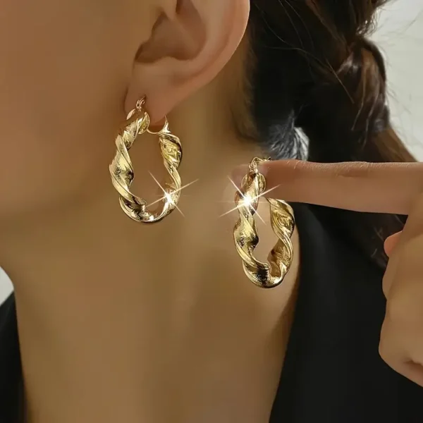 A close-up captures a person wearing Twisted Rope Golden Hoops Earrings - Elegant Copper Gift. One of the earrings is gently touched by a finger, causing it to sparkle under the light. The individual has dark hair pulled back and is dressed in a black garment.