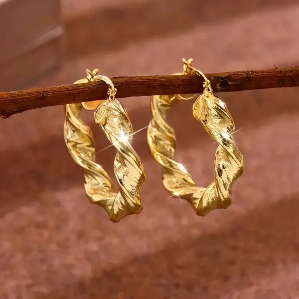 A pair of Twisted Rope Golden Hoops Earrings- Elegant Copper Gift hang from a thin branch. The golden hoop earrings feature a shiny, textured surface that catches the light, creating a sparkling effect. The background is a soft brown fabric.