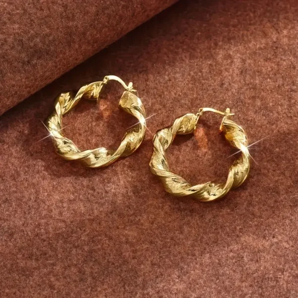 A pair of Twisted Rope Golden Hoops Earrings- Elegant Copper Gift resting on a brown textured surface. These earrings feature a shiny, polished finish with intricate detailing that adds to their elegance. The lighting highlights the gold's reflective quality, giving a sparkling effect.