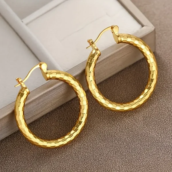 The 14K Gold Plated Delicate Hoop Earrings - Vintage Bohemian Style, featuring a textured, faceted design and a shiny, reflective surface, rest on a brown surface next to a wooden jewelry box. These elegant earrings include a latch-back closure for secure wear.