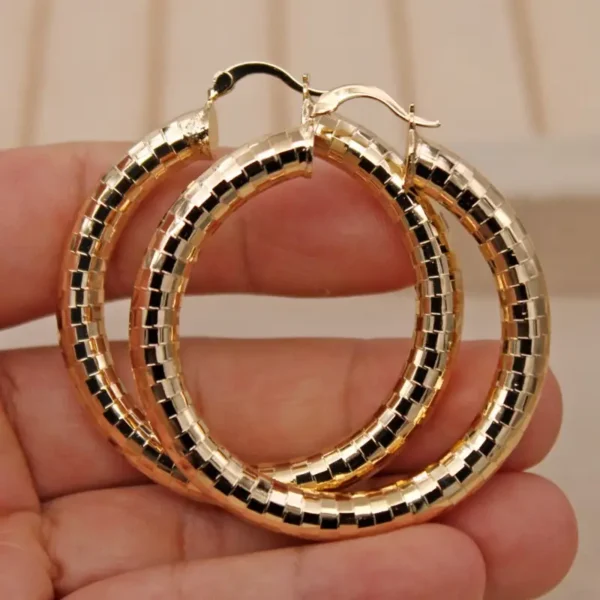 A hand holding a pair of Elegant Copper Oval Hoop Earrings, perfect for weddings and celebrations, showcases their intricate segmented design as they reflect light. The shiny metallic finish of the hoops stands out against a blurred background featuring neutral tones.