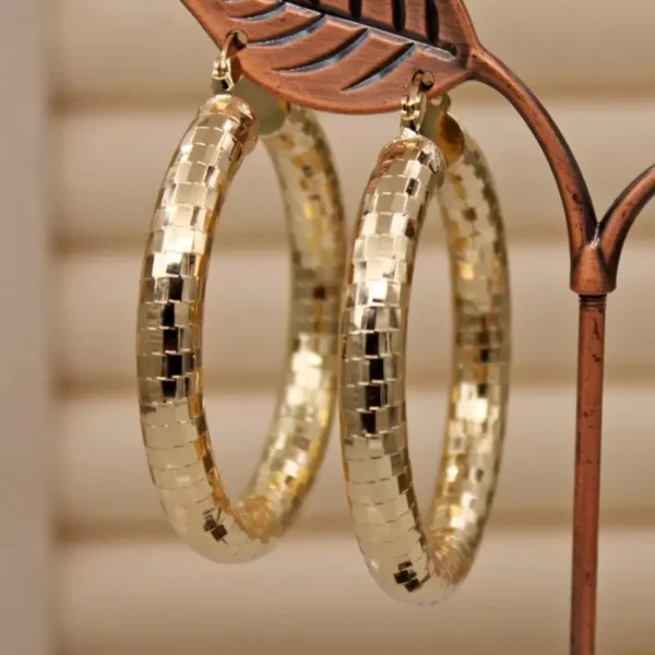 A pair of Elegant Copper Oval Hoop Earrings, perfect for weddings and celebrations, hang from a decorative metal stand. The large earrings feature a checkered texture and have a glossy finish that reflects light beautifully against a neutral, blurred background.