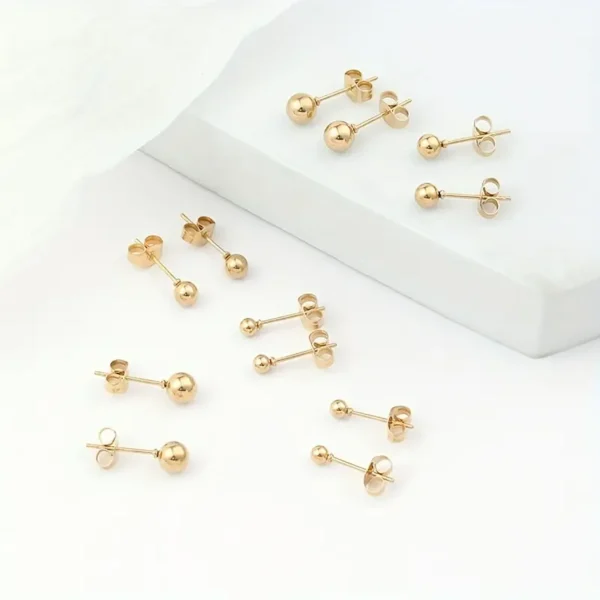 A selection of Golden Ball Stud Earrings: Punk Style, Everyday Stainless Steel is beautifully displayed on a white surface. These minimalist gold earrings feature slight variations in design and size, showcasing larger spherical ends on some pairs while others have smaller, more refined ends.