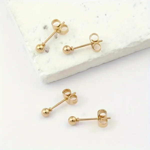 A set of four Golden Ball Stud Earrings: Punk Style, Everyday Stainless Steel is displayed on a textured white stone surface. The earrings are arranged with one pair on the stone and the other pair next to it. Each earring features a small gold ball at the end of a stainless steel post with butterfly backings.