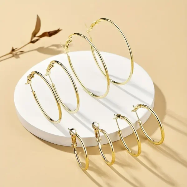 The 4 Pair Hoop Earrings Set: Minimalist, Stainless Steel, Everyday Chic is elegantly arranged on a round, white platform against a beige background. The set features two pairs of gold hoops and one pair of silver hoops, each beautifully displayed with a small dried branch nearby.