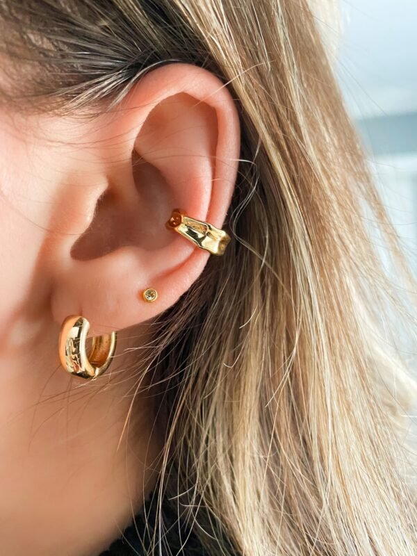 Close-up of a person's ear, featuring the Chunky Small 14kt Gold Hoops Earrings on the cartilage. Additionally, there's a small stud on the lobe and a small gold creole in the tragus piercing. The person's light brown hair partially covers the top of their ear.