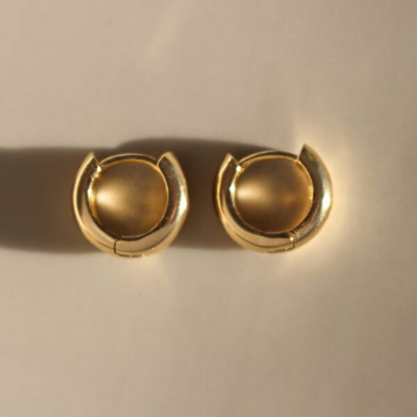 A pair of Chunky Small 14kt Gold Hoops Earrings with a spherical design are placed on a smooth, light-colored surface. The gold hoops feature a slightly raised, domed center within their circular structure. Shadows cast by the earrings are visible underneath them, adding depth to these small gold creoles.