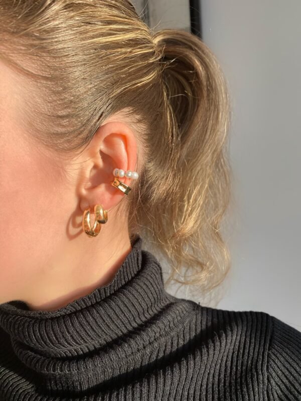 A close-up of a person’s ear adorned with multiple earrings. Three gold hoop earrings, a ring with three small pearls, and Chunky Small 14kt Gold Hoops Earrings are worn together. The person has blonde hair tied back and is wearing a black ribbed turtleneck. The background is neutral and softly lit.