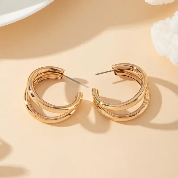 A pair of Chunky C-Shaped Double Hoop Earrings rests on a beige surface. Each earring features two interwoven bands, creating a stylish and elegant design. The minimalist style jewelry casts soft shadows, enhancing the overall aesthetic appeal.