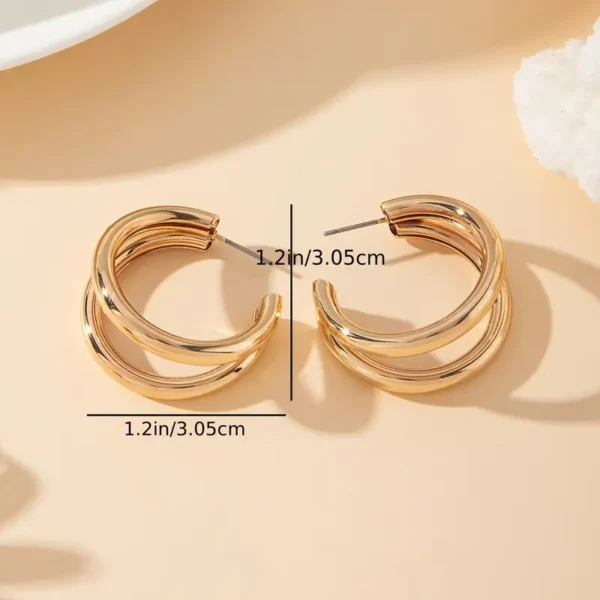 Displayed on a beige surface, the Chunky C-Shaped Double Hoop Earrings showcase their gold-tone, double hoop design. The earrings are laid flat, revealing dimensions of 1.2 inches (3.05 cm) in both height and width. The shadows cast create a soft, minimalist aesthetic.