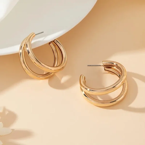 A pair of Chunky C-Shaped Double Hoop Earrings is displayed against a neutral, beige background. One earring rests against a white surface, while the other lies flat beside it, highlighting their elegant gold design and reflective finish.