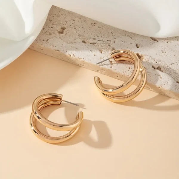 A pair of Chunky C-Shaped Double Hoop Earrings is elegantly displayed on a neutral, textured surface. The earrings feature a shiny, polished finish, and their curved design casts subtle shadows, enhancing their sophisticated appearance. White fabric is draped softly in the background, adding to the minimalist style jewelry aesthetic.