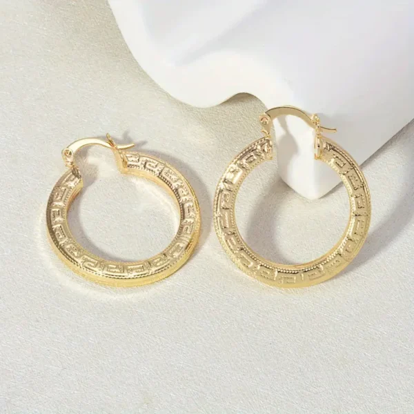 A pair of Elegant 14K Gold Plated Hoop Earrings with Great Wall Print lies on a light-colored surface. The earrings feature an intricate pattern reminiscent of the Great Wall etched along the outer edge, adding a touch of elegance. One earring is slightly propped up against a white sculptural object.