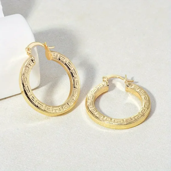 The Elegant 14K Gold Plated Hoop Earrings with Great Wall Print, adorned with intricate engravings and a geometric pattern, rest on a light beige surface. The earrings are displayed both standing and lying flat.