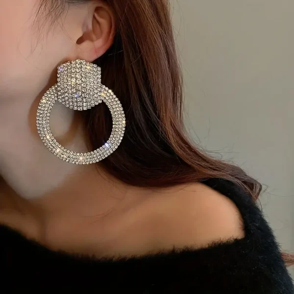 A close-up of a person's ear adorned with Elegant Rhinestone Hollow Circle Dangle Earrings. The earring features a glittering hoop design with a diamond-encrusted circular stud at the top. The person is wearing a black, off-the-shoulder top, and the background is blurred and neutral.