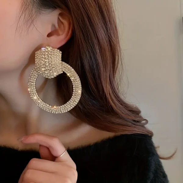 A close-up of a person's ear adorned with large, glittering Elegant Rhinestone Hollow Circle Dangle Earrings. The earrings are made of sparkling, rhinestone-encrusted material. The person is wearing a black top and has long, brown hair partially covering the ear. Their hand rests near the earring, showing a thin silver ring.