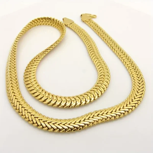 A pair of intricately designed gold chains lie on a light surface. One chain is slightly longer than the other, and both feature a herringbone pattern that reflects light, giving them a polished and elegant appearance. The Hip Hop Royalty: Gold Plated Herringbone Set embodies the essence of hip hop jewelry with its striking design.