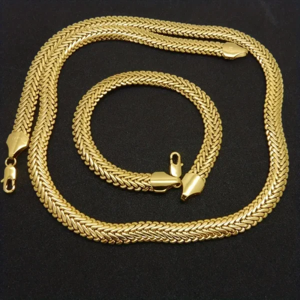 The Hip Hop Royalty: Gold Plated Herringbone Set, featuring a gold-plated necklace and bracelet, is displayed on a black surface. Both pieces have a shiny, polished appearance and feature clasp closures. The intricate herringbone chain design and bright gold color stand out against the dark background, making it perfect as hip hop jewelry.