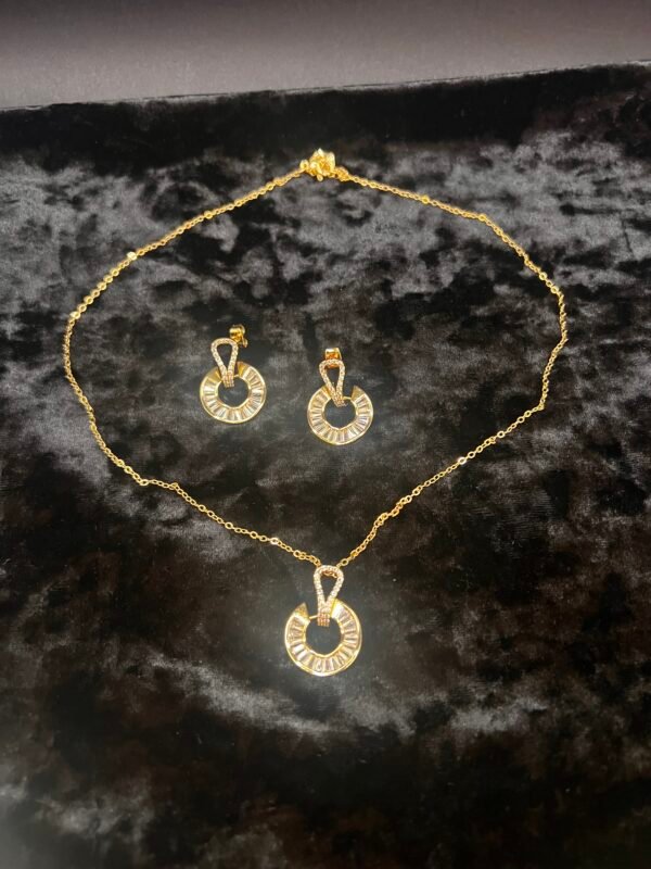 A 3-piece gold-plated jewelry set, crafted from stainless steel and adorned with cubic zirconia, displayed on a black velvet surface. The gold necklace and matching earrings feature circular pendants with a unique geometric design, creating an elegant and cohesive look. The delicate chain and earring hooks emphasize the intricate details.