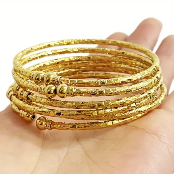 A hand is holding several stacked pieces from the 6-Piece African Bead Bracelet Set - Luxurious Bridal Gift, Ethiopian Inspired. The thin gold bangles are adorned with small spherical beads and intricate textured patterns, resembling a traditional Egyptian jewelry set. The plain white background highlights the shiny and detailed design of this elegant jewelry collection.